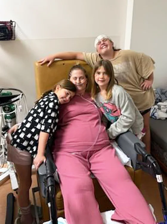 Angela Goodrich sitting in a chair wearing pink clothing surrounded by three of her children hugging her, showing family support during her cancer journey.