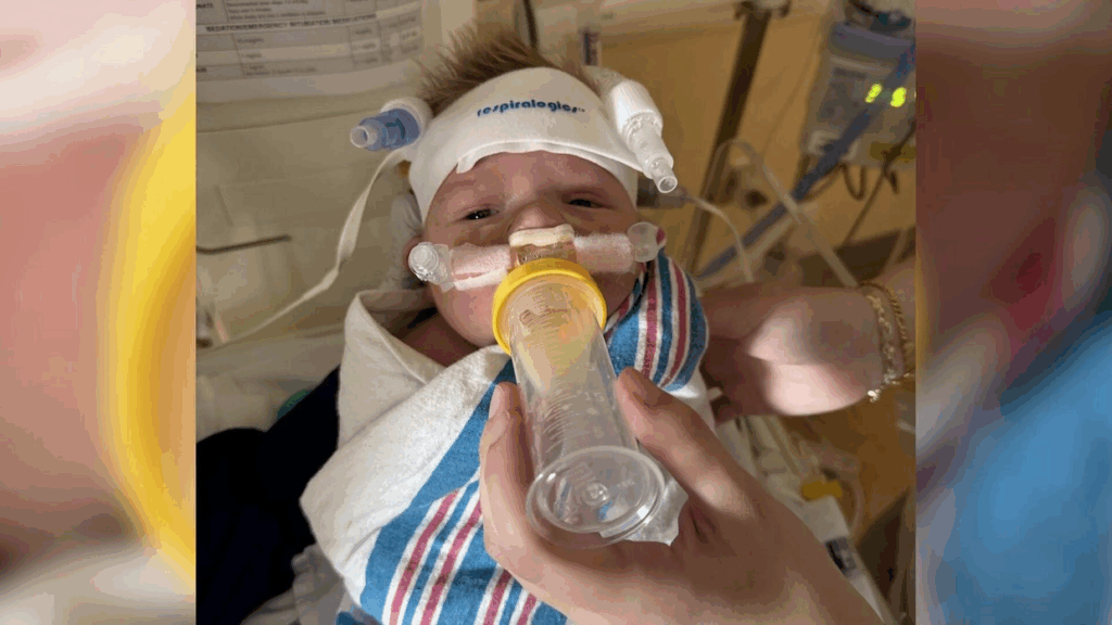 Baby Cole in hospital with critical care monitoring equipment, wearing a white cap labeled "Inspirologist" and receiving oxygen support through a nasal tube. A caregiver's hand is holding a bottle to feed Cole, who is wrapped in a striped hospital blanket following his severe bleeding incident after circumcision.