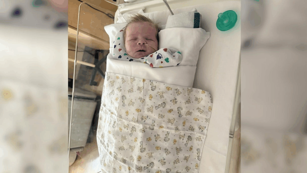 Baby Cole resting in his hospital bassinet, covered with a patterned blanket featuring baby animals. Medical equipment is visible in the blurred background, showing the clinical setting where Cole receives ongoing care for complications from his blood loss after the procedure.