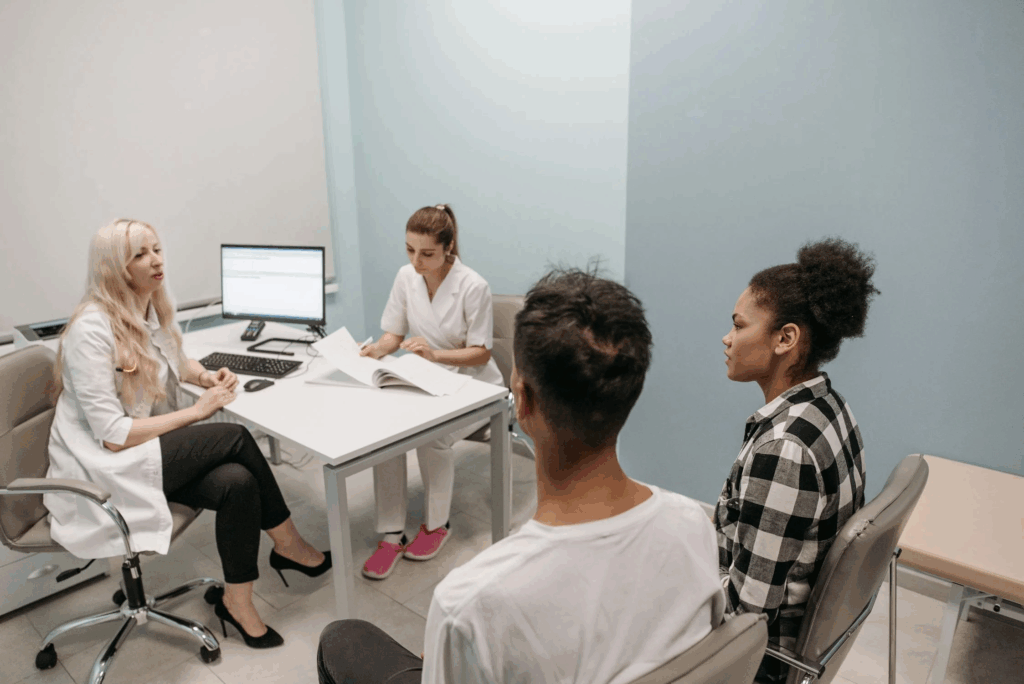 Medical consultation taking place in a clinical setting with healthcare providers in white coats discussing patient information with two people seated across from them at a desk with a computer monitor, paperwork visible on the table.