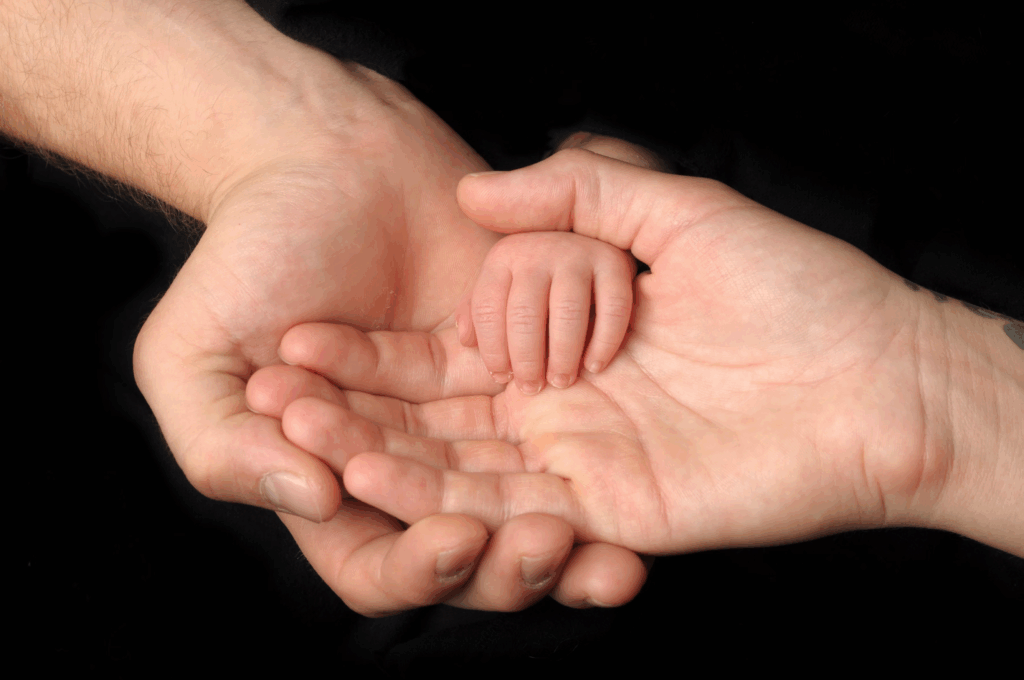 Close-up of an adult's hands gently cradling a tiny infant's hand, showing the dramatic size difference and conveying parental care and protection against a black background.