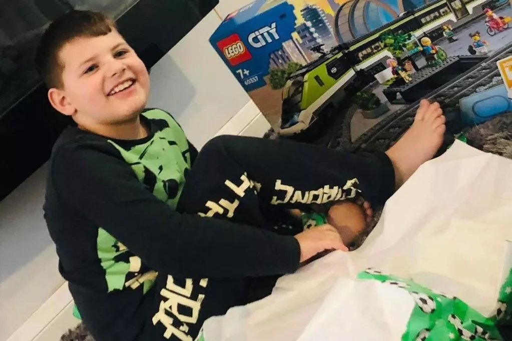 Joshua, the 8-year-old boy who tragically lost his life, smiling while sitting with LEGO City toys. He's wearing black and green pajamas with text on them, looking happy as he plays with his building sets, before falling victim to helium asphyxiation that struck without warning.