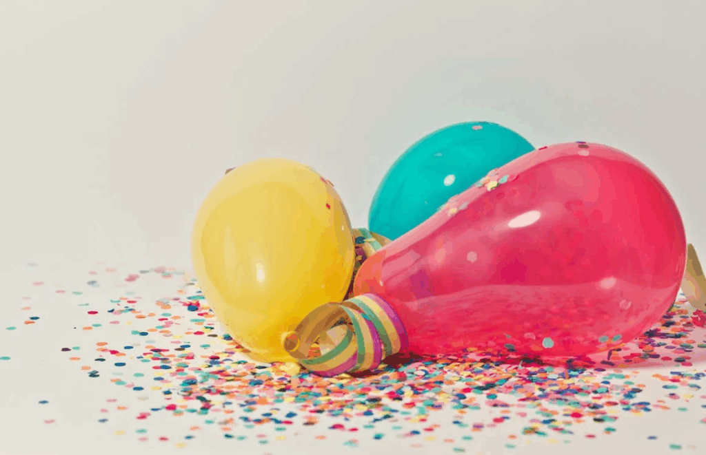 Colorful party balloons - red, teal, and yellow - resting on a surface scattered with confetti. The deflating balloons represent a hidden danger at celebrations that can lead to tragic helium-related deaths when children play with them unsupervised.