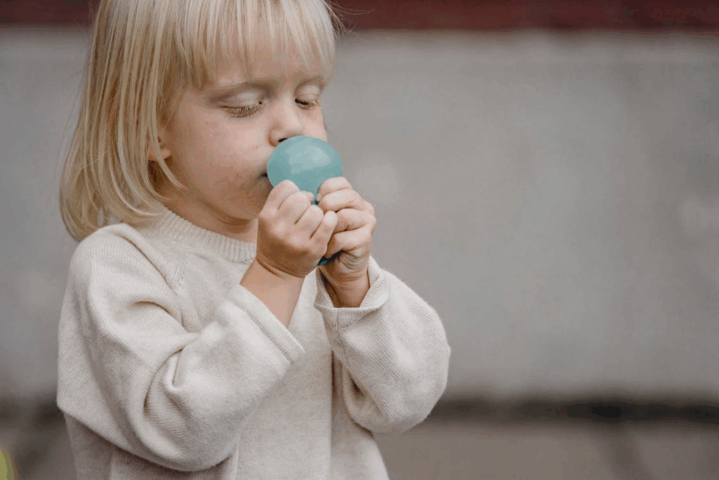 A young child with blonde hair and bangs blowing up a small blue balloon, demonstrating how children innocently interact with balloons, not recognizing the serious risks they can pose when filled with helium instead of air.