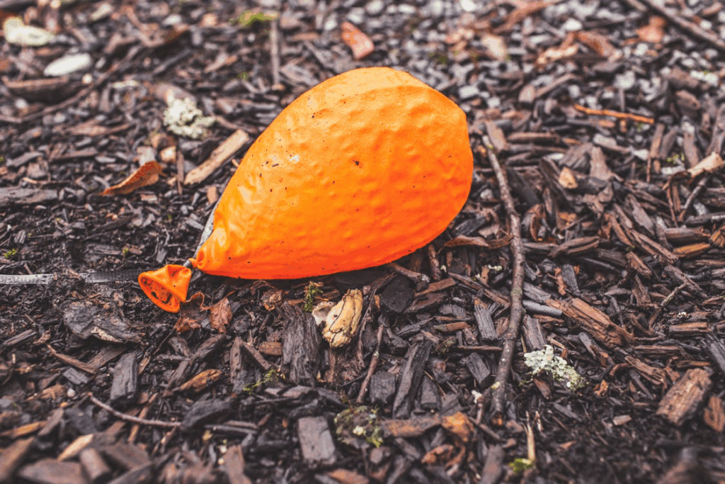 A deflated orange balloon resting on dark mulch ground, representing the dangers that remain even after parties end, when partially deflated helium balloons can still pose significant suffocation risks to curious children.