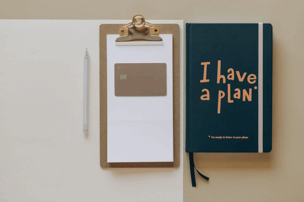 A clipboard with a blank sheet of paper and a pen is placed next to a notebook labeled "I have a plan." The setup suggests planning and organization, emphasizing the importance of creating structured financial frameworks when supporting adult kids.