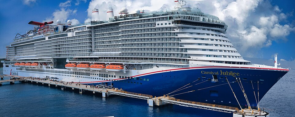 A full side view of the massive Carnival Cruise ship Jubilee docked at port. The vessel features the distinctive Carnival Cruise line design with a navy blue hull adorned with the 'Carnival Jubilee' name, white superstructure with multiple rows of cabin balconies, and red funnel with the Carnival logo. The ship is connected to the dock by gangways, with blue ocean waters surrounding it and a partly cloudy sky overhead.