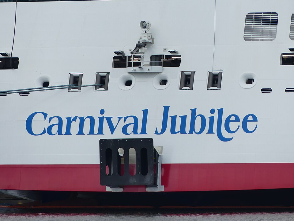 Close-up view of a cruise ship hull, showing the ship's name emblazoned in blue script lettering against the white hull. The vessel features a distinctive red stripe along the bottom edge and several windows and portholes are visible. Security cameras and mounting equipment can be seen on the upper portion of the image.