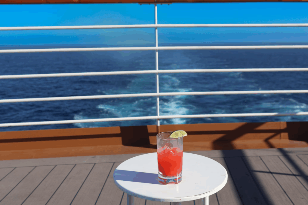 A red fruity cocktail with a lime garnish sits on a small white round table on the deck of a cruise ship. Through the railings in the background, the deep blue ocean stretches to the horizon with the ship's wake visible in the water. The wooden deck floor shows shadows cast by bright sunlight, creating a typical relaxing cruise atmosphere.