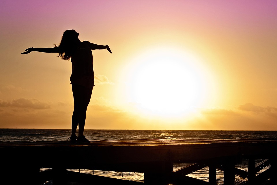 Silhouette of a person with arms outstretched facing a sunset over the ocean, representing freedom, healing, and new beginnings.