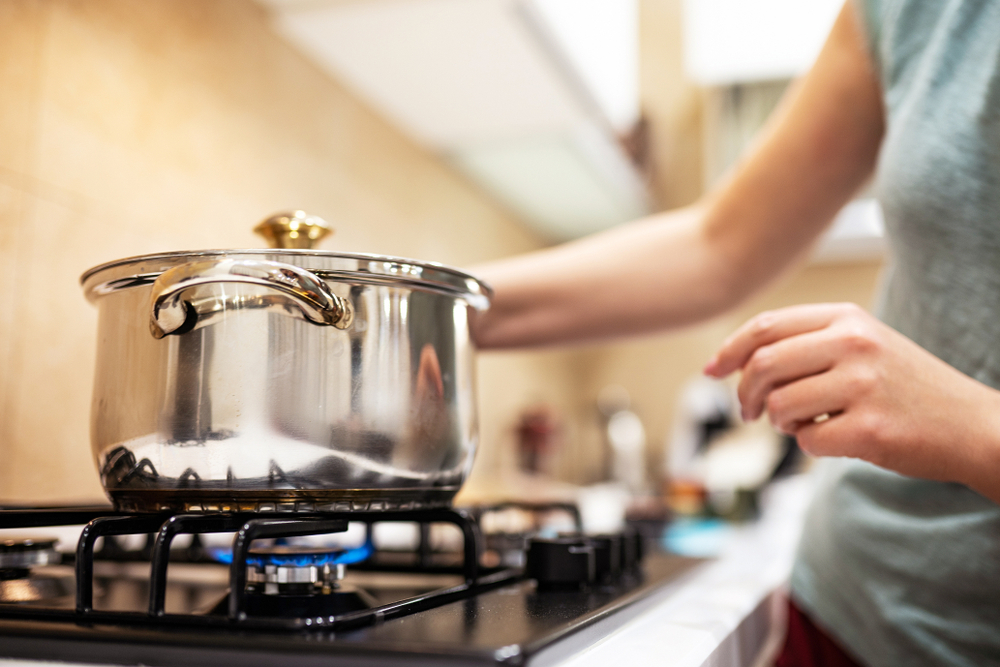 Beautiful young woman housewife prepairing dinner, hold in hands big steel saucepan, standing it on gas-stove.