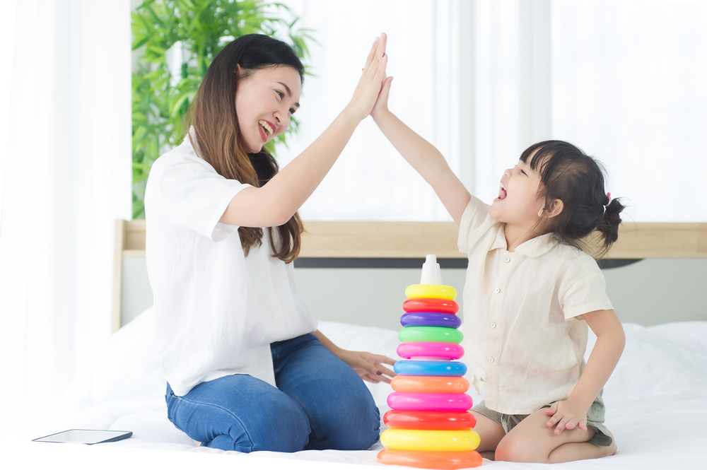 Happiness mother and daughter sit on a bed in the house doing high five while playing the colorful toy together, family joyful together in the house on holiday