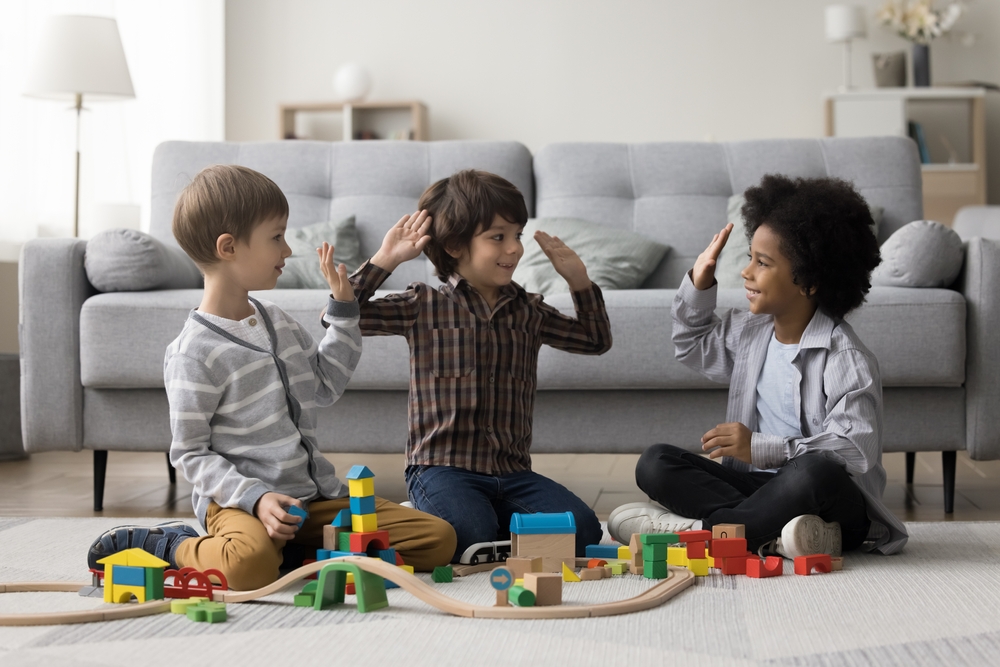 Diverse team of cheerful kids playing game on home floor, constructing towers, city road from toy building bricks, giving group high five, celebrating teamwork success
