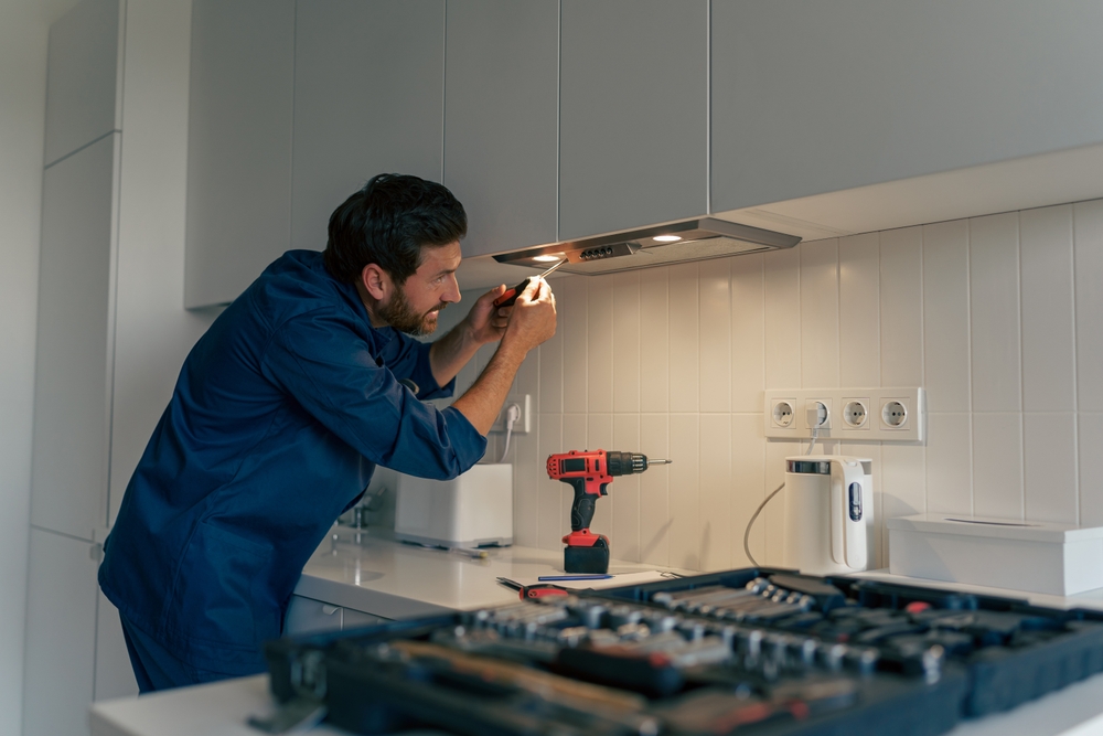 Professional male worker in uniform repairing modern cooker hood in kitchen
