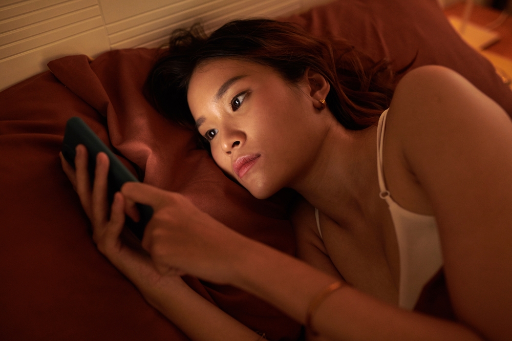 Young woman doomscrolling social media on smartphone before sleep