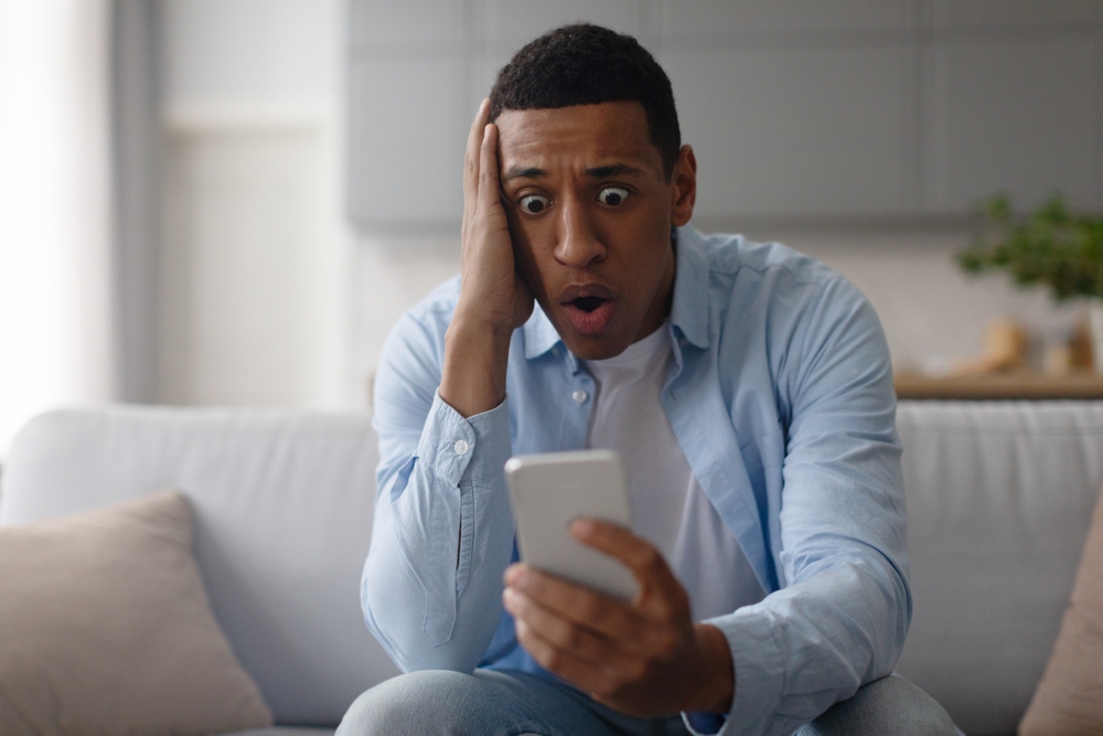 Shocked black man dealing with cellphone issue, looking at phone, reading unpleasant news or message, sitting on sofa at home and touching head