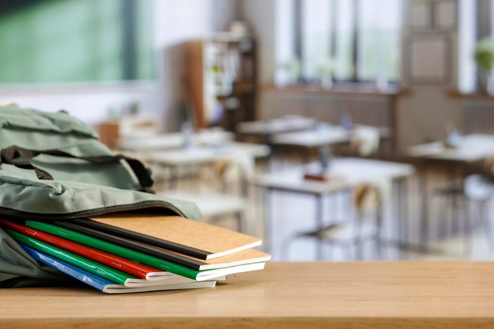School backpack and school stuff with space for products on wooden desk in classroom interior. Chalkboard wall background. Back to school. Copy space for school supplies and accessories.