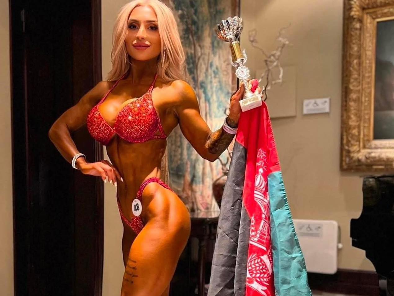 Roya Karimi posing in her competition outfit while holding an Afghan flag and her award she won.