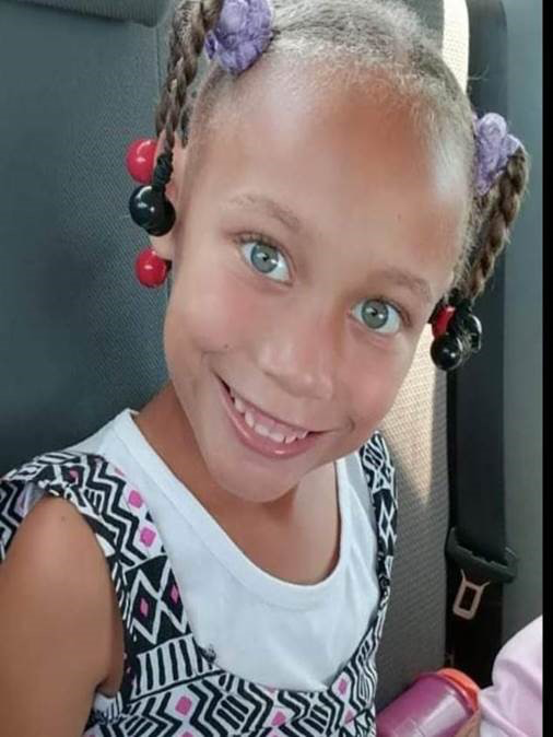 Joshlin Smith, a 6-year-old girl with braided hair and colorful beaded accessories, smiling at the camera while wearing a black and white patterned dress.
