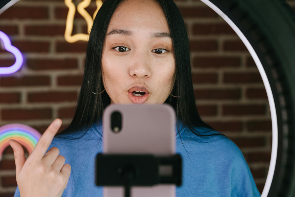 Content creator speaking into smartphone while filming, with ring light and neon signs in background
