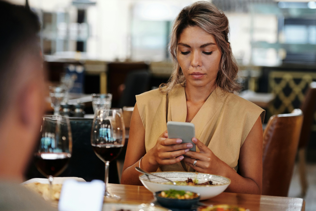 Woman distracted by phone during dinner date, illustrating how technology creates barriers to meaningful conversation between partners.