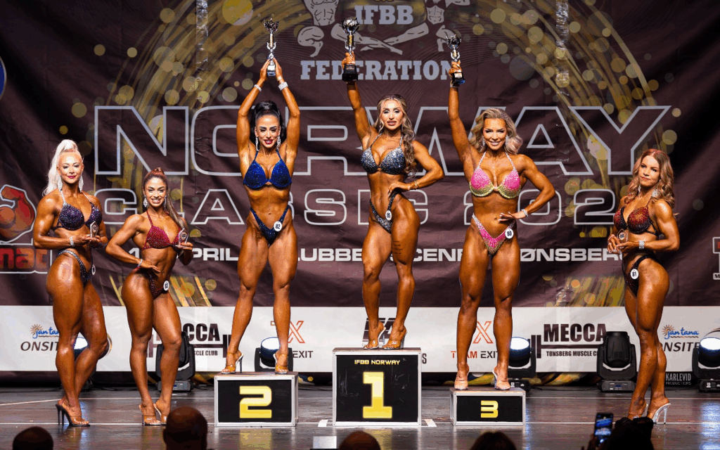 Roya Karimi on stage with competitors, holding her gold metal in the air at the IFBB Norway competition 2024.