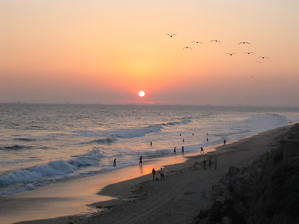 Huntington Beach sunset with silhouettes of people walking along the shoreline as birds fly overhead, showcasing the coastal lifestyle that delivers happiness.