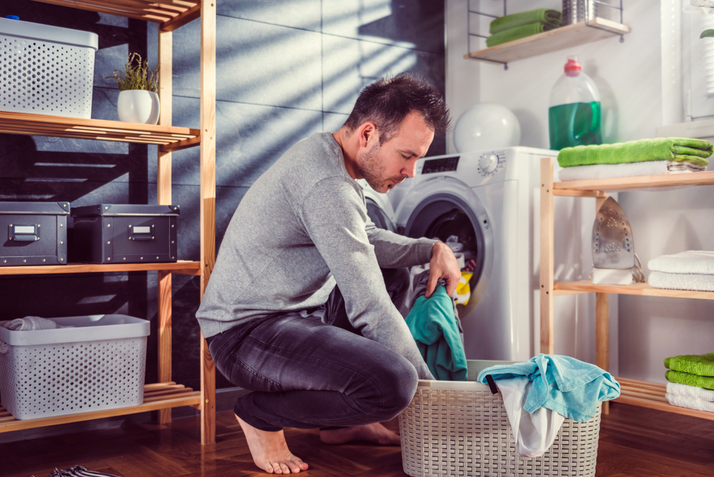 Man wearing grey sweater putting clothes into washing machine at laundry room
