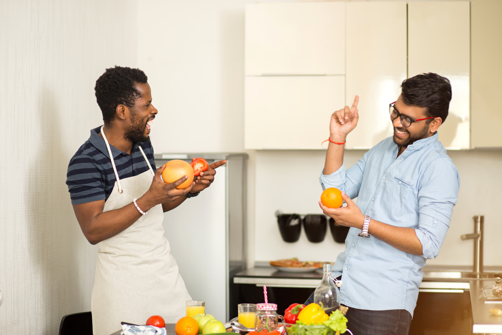 Cheerful indian young man in glasses and african american man wearing apron having fun in kitchen. Two male friends standing near desk with fruits and vegetables and laughing while cooking breakfast.