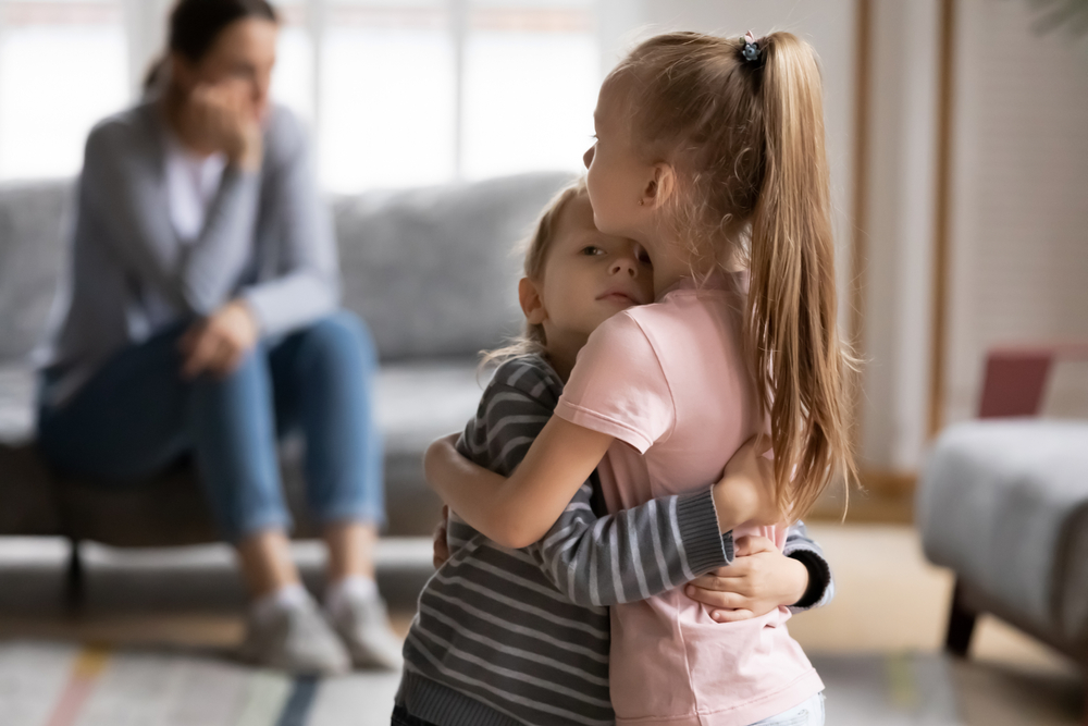 Empathic elder sister cuddling comforting little brother, while stressed annoyed mother sitting on couch at home. Small children siblings supporting each other after family quarrel with mum.