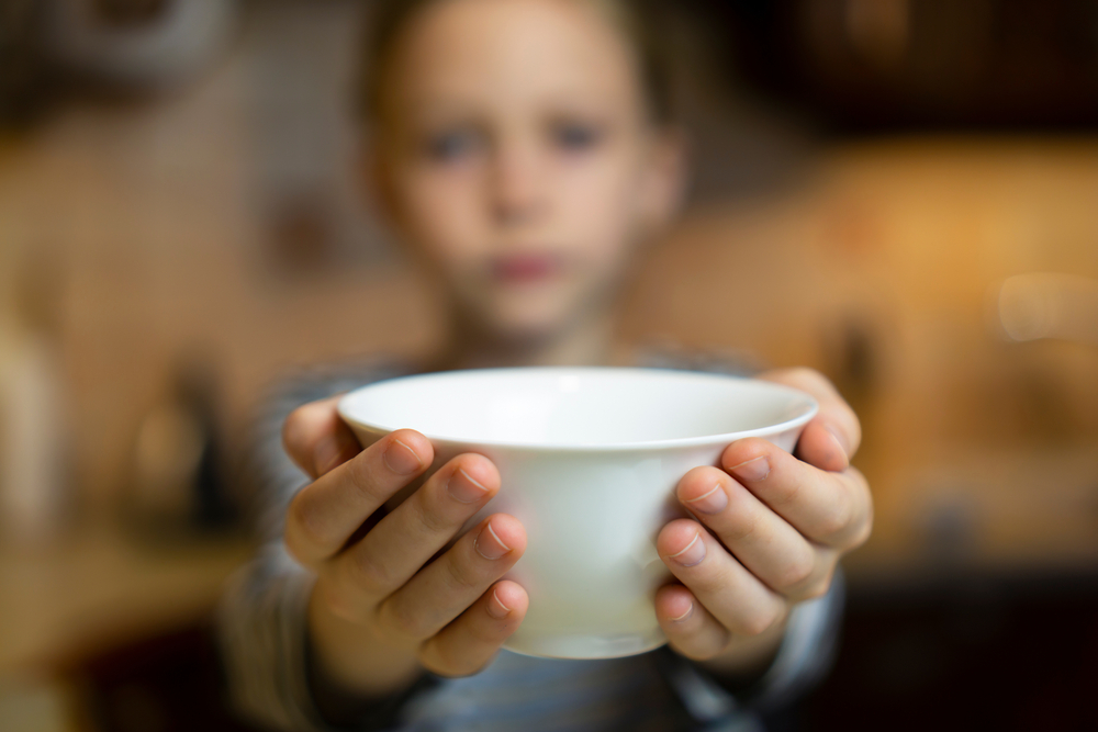 Unrecognizable child girl reaching out hands holding white empty bowl plate offering food or asking for food. Shallow focus. Giving concept. Hungry children