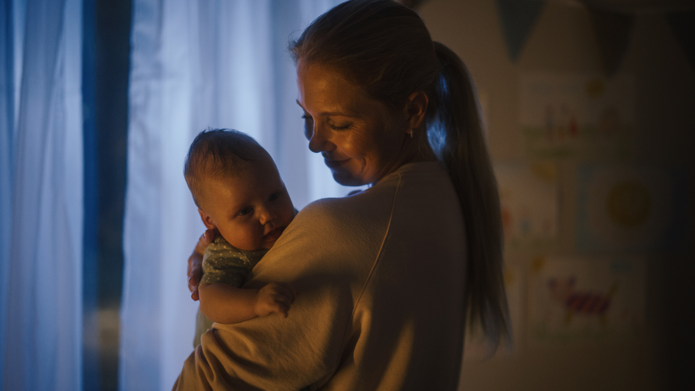 Happy Mother Holding Cute Newborn Baby in Cozy Dark Kids Bedroom in the Evening. Caring Mom Hugging the Neonate Toddler and They are Bonding Before Sleep. Concept of Childhood, New Llife, Parenthood.
