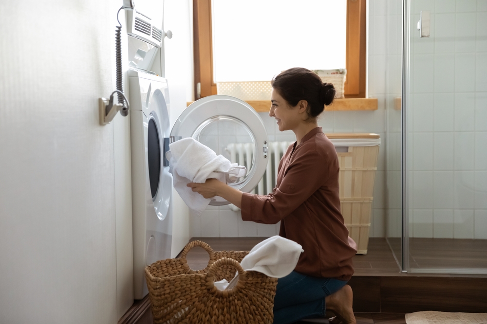 Young happy Indian housewife doing housework putting clothes into washing machine in cozy domestic laundry room. Routine house work, housekeeping, washer-dryer modern appliance or detergent ad concept
