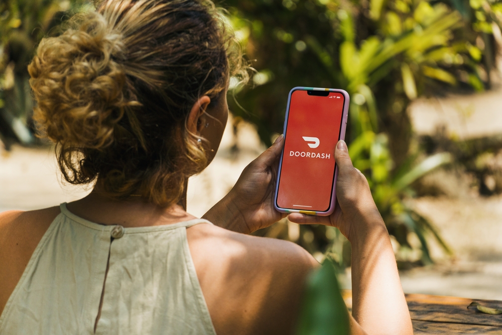 Girl in the park holding a smartphone with Doordash online food ordering and delivery platform app on the screen. Rustic wooden table. Rio de Janeiro, RJ, Brazil. January 2022.