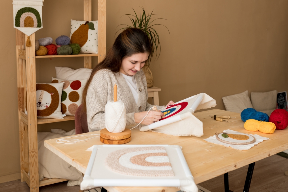 Punch needle embroidery. Young woman pushing the punchneedle straight down into the foundation fabric in workshop. Female embroidering handmade picture rainbow on canvas. Handicraft, hobby, diy