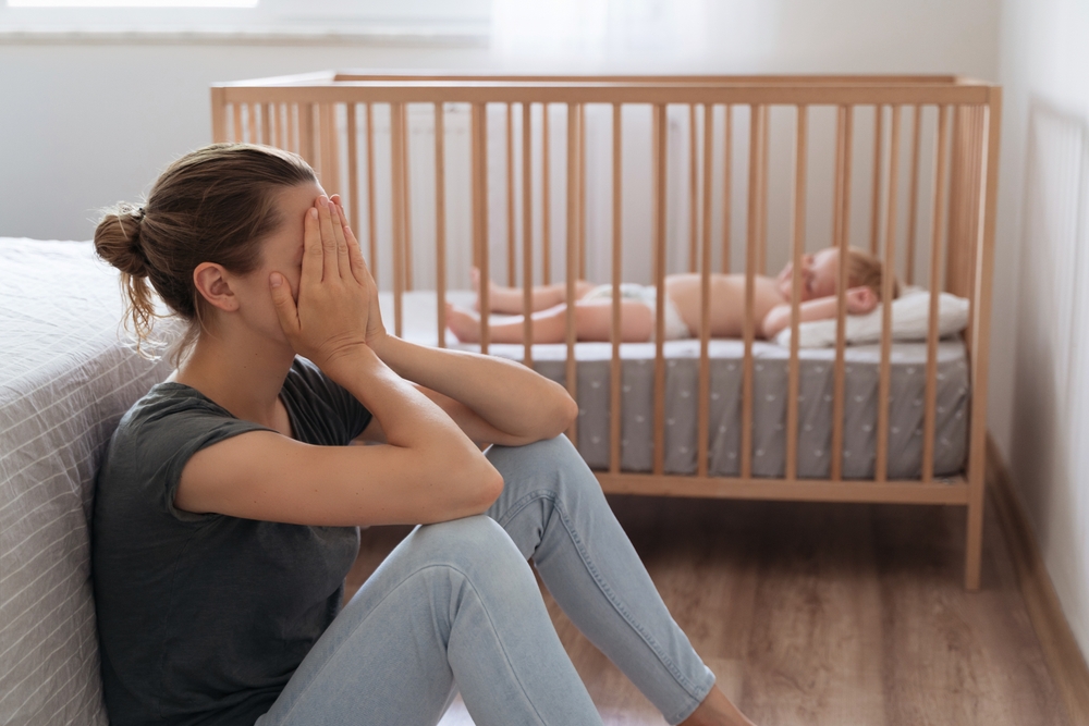 Side view of mother covering face crying from exhaustion sitting on floor next to baby crib while child napping, can't handle difficulties of maternity, having postnatal depression symptoms
