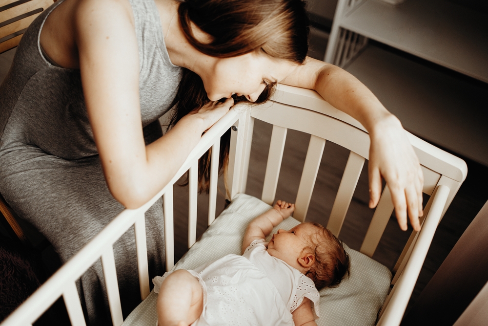 The child sleeps in a crib, the mother sits next to her and tenderly looks at her daughter
