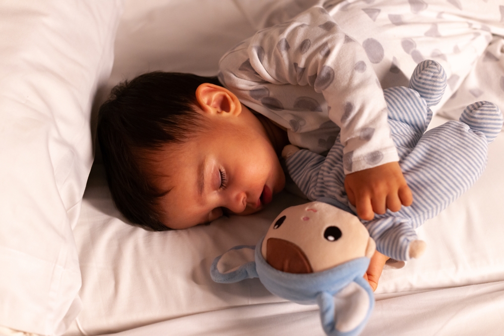 Baby sleeping with mouth open and cuddling teddy bear in bed
