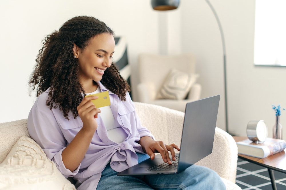 Online shopping. Positive hispanic or brazilian curly haired woman, using credit card and laptop to buying something, paying online making purchase in online store while sitting at home, smiling