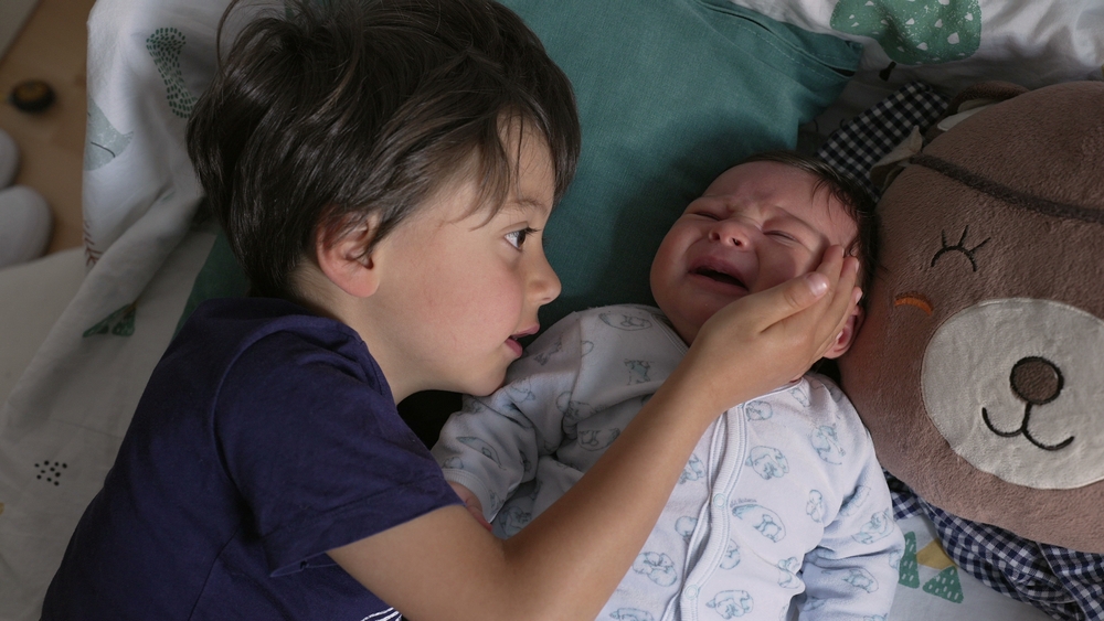 Young boy gently comforting his crying newborn sibling, both lying on a bed with a plush pillow, showcasing a tender sibling bond in a peaceful home setting