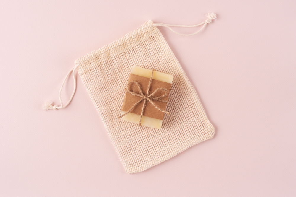 Handmade natural soap bar wrapped in kraft paper with twine bow, placed on a reusable cotton mesh bag against a soft pink background. Perfect for eco-friendly, sustainable, and skincare concepts.
