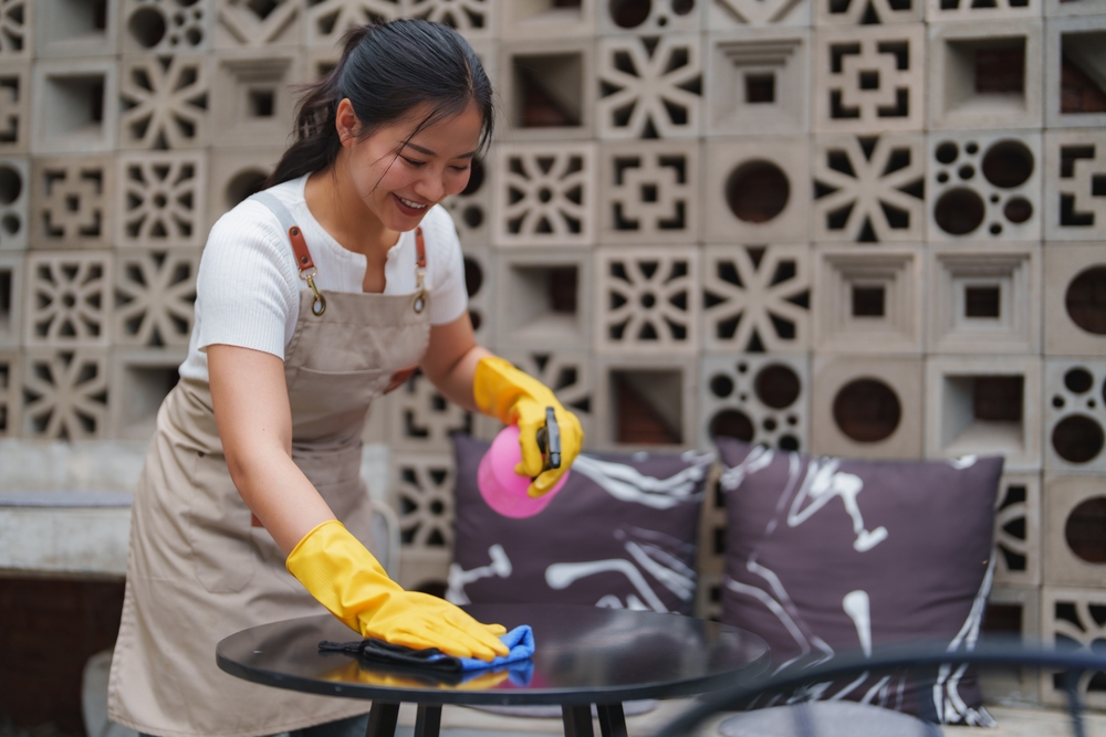 Young smiling waitress wearing yellow gloves and apron cleaning table with disinfectant spray and blue cloth, preparing outdoor cafe for opening