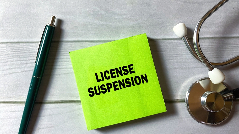 The concept of License Suspension is written on a sticky note with a stethoscope isolated on a wooden background.