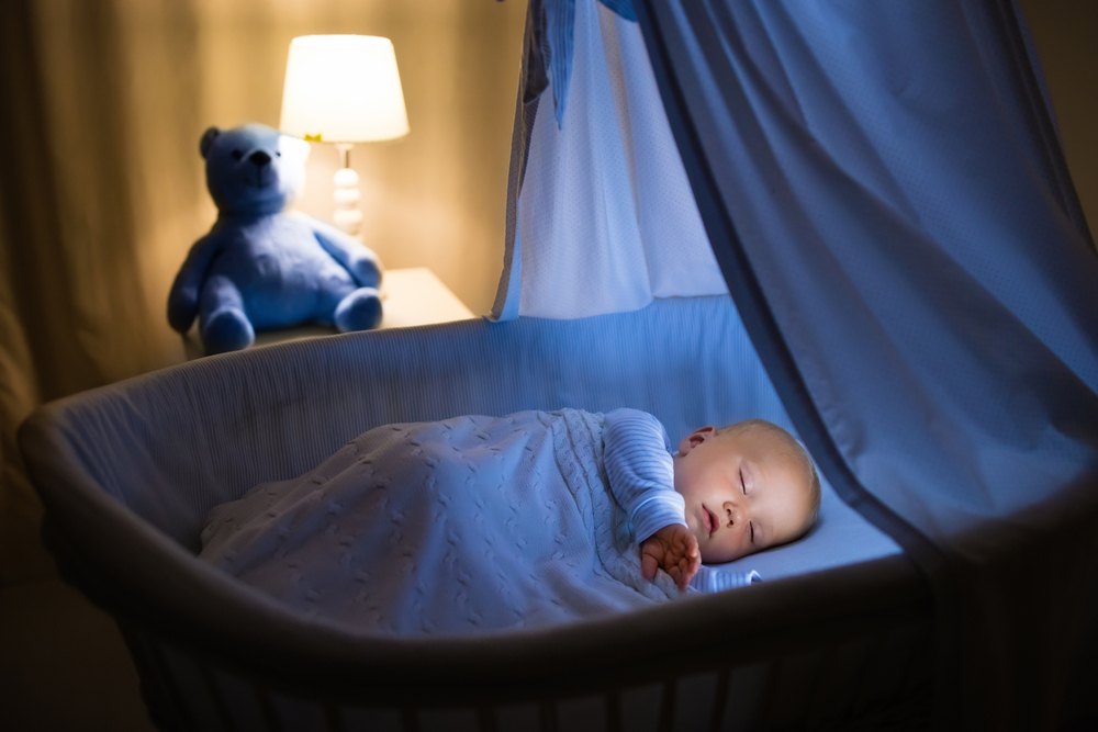 Adorable baby drinking milk in blue bassinet with canopy at night. Little boy in pajamas with formula bottle getting ready to sleep in dark room with crib, lamp and toy bear. Bed time drink for kids
