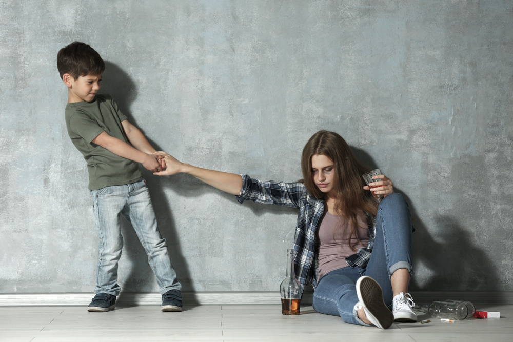 Woman drinking alcohol while little son pulling her hand near grey wall
