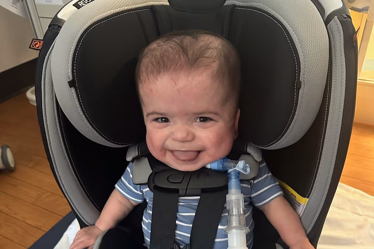 Barrett Brasfield grinning in his car seat with a tracheostomy tube visible, ready to go home for the first time.