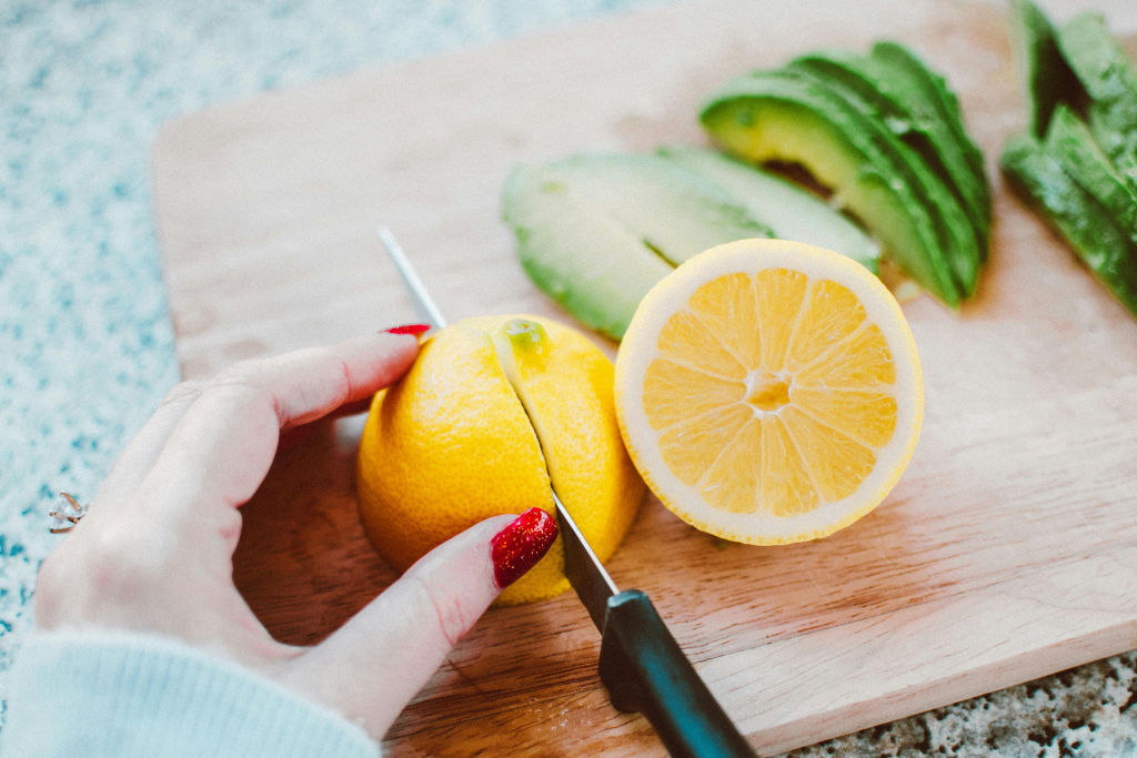 Hands slicing fresh orange on a wooden cutting board, with lemon halves and avocado nearby.