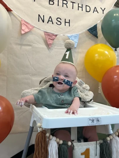 Nash Keen celebrates his first birthday sitting in a high chair, wearing a green party hat and his hearing aids, surrounded by colorful balloons and birthday bunting that reads "BIRTHDAY NASH." He appears happy and engaged during this milestone celebration.