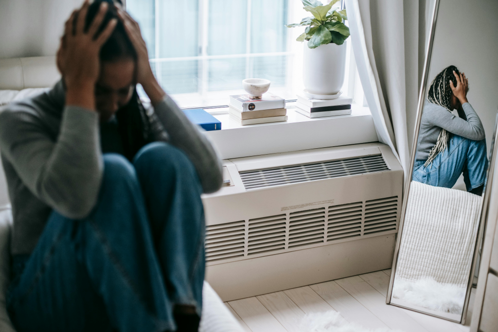 Person in distress sitting by a window, on a bed, with their troubled reflection visible in a nearby mirror.