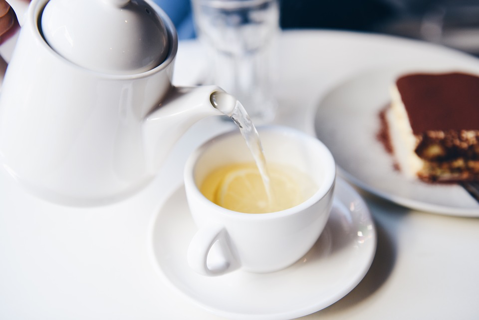 Steaming herbal tea being poured into a white cup, with cake visible in the background. showcasing everyday uses for lemons in cooking and wellness.